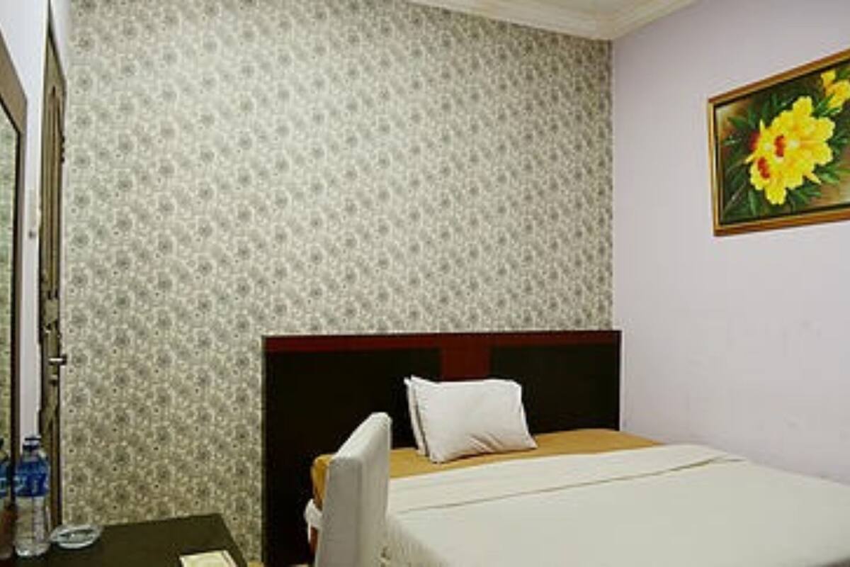 A hotel room with a patterned wallpaper, a bed with a wooden headboard, a white pillow, and a painting of flowers on the wall.