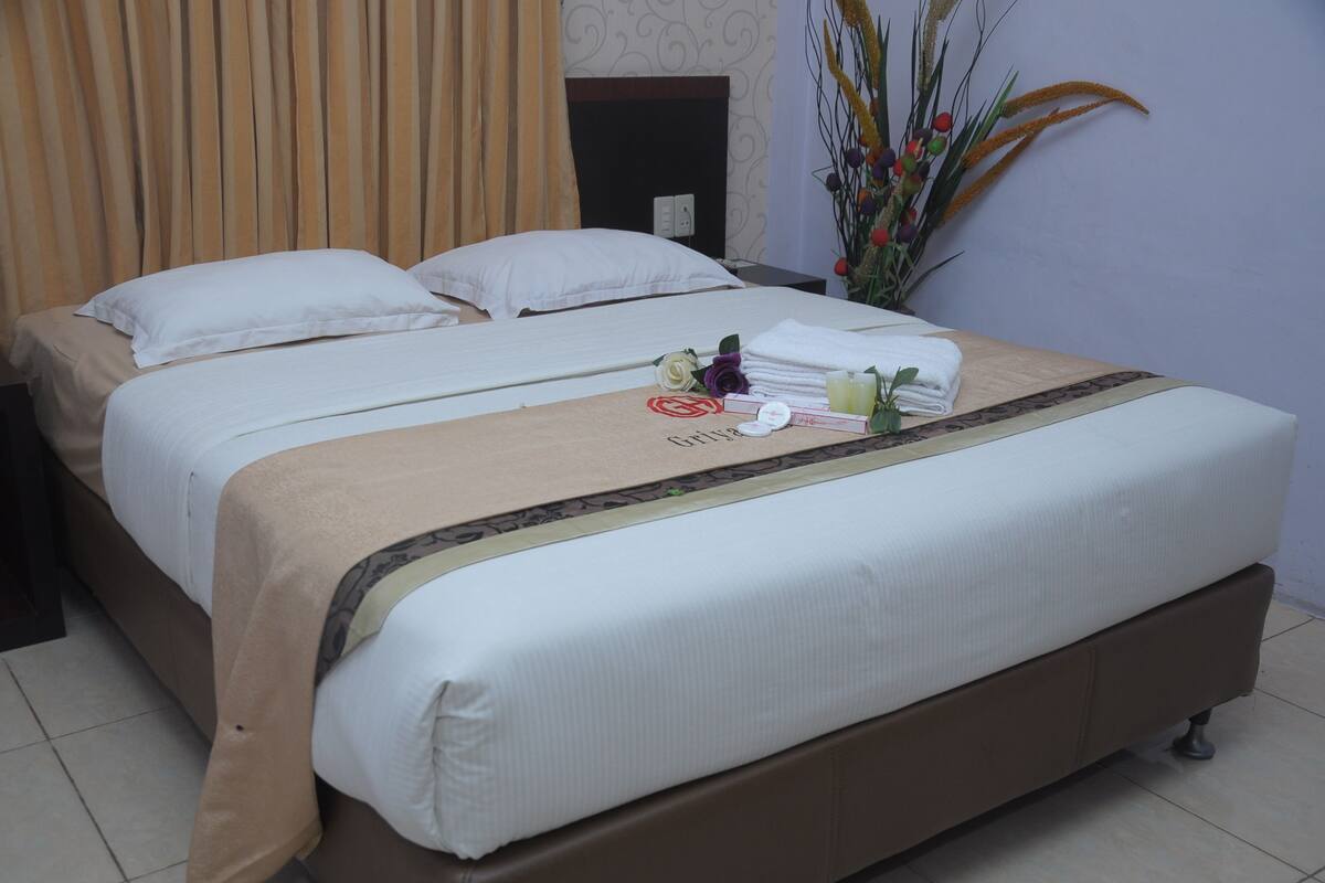 A double bed with a beige bedspread and white pillows, a small table with toiletries, and a decorative plant.