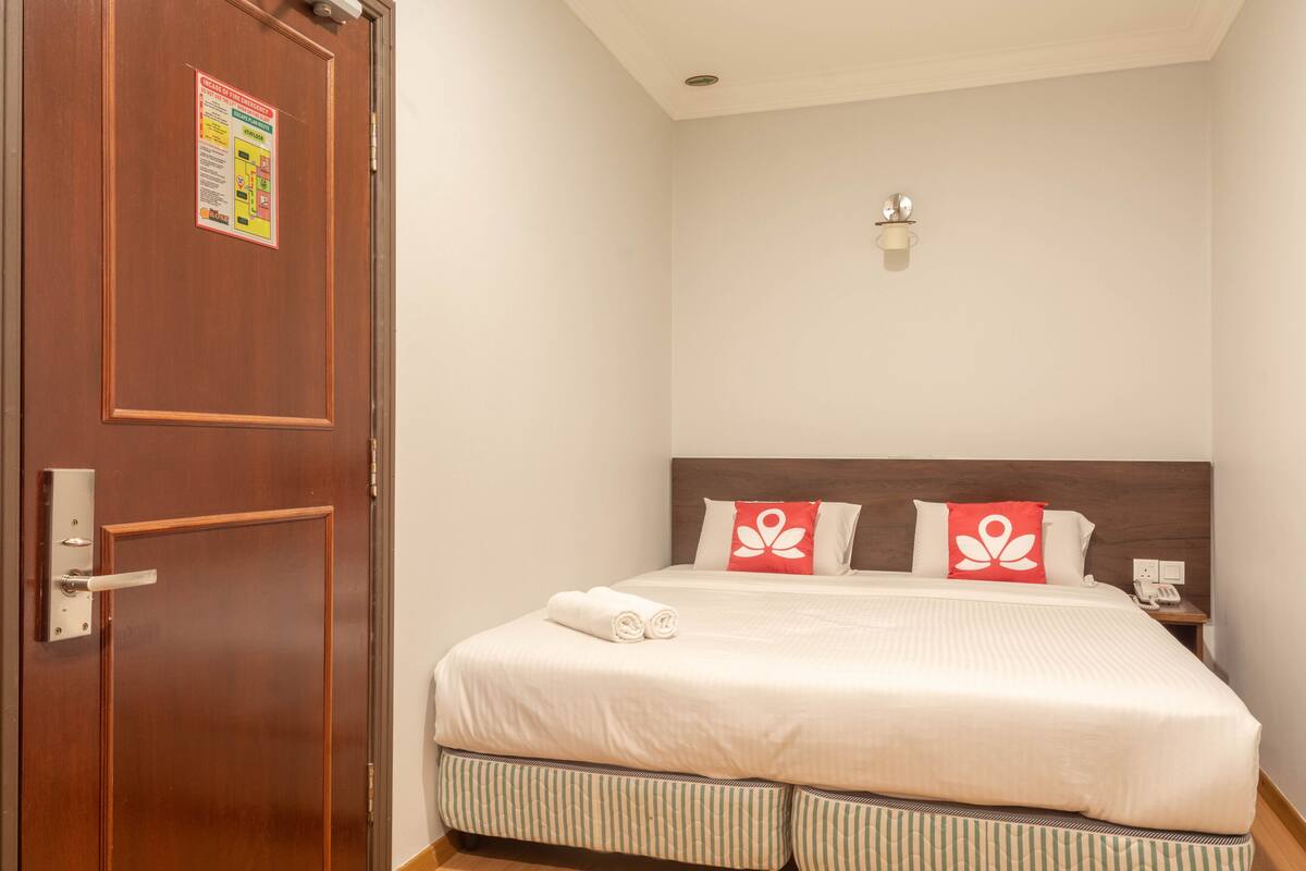 A hotel room with a wooden door, a bed with white bedding and two red pillows, a bedside table with a phone, and a wall-mounted light fixture.