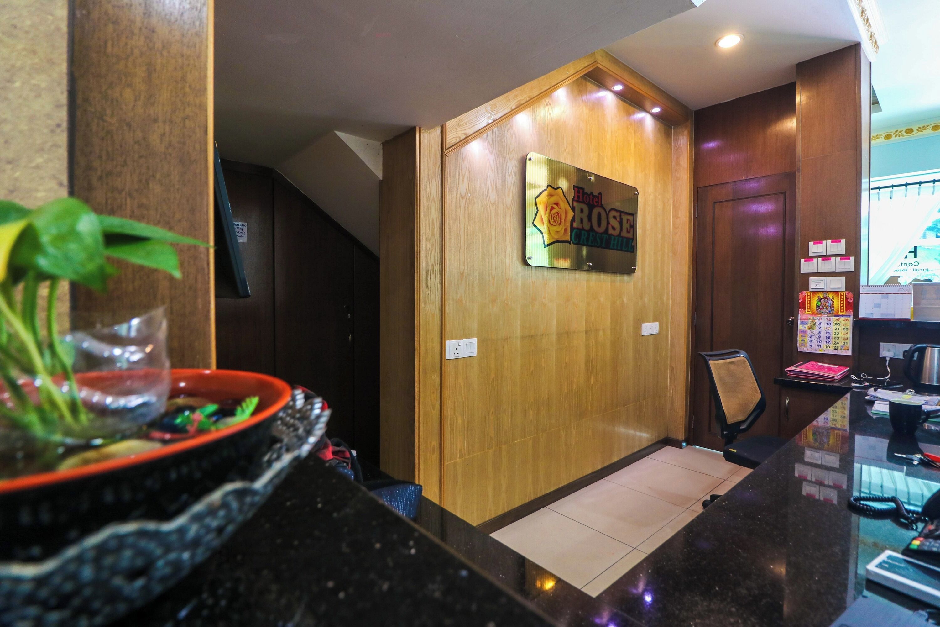 A reception desk with a wooden panel wall, a sign reading "Hotel Rosle Crest Hill," and a plant in a black bowl.