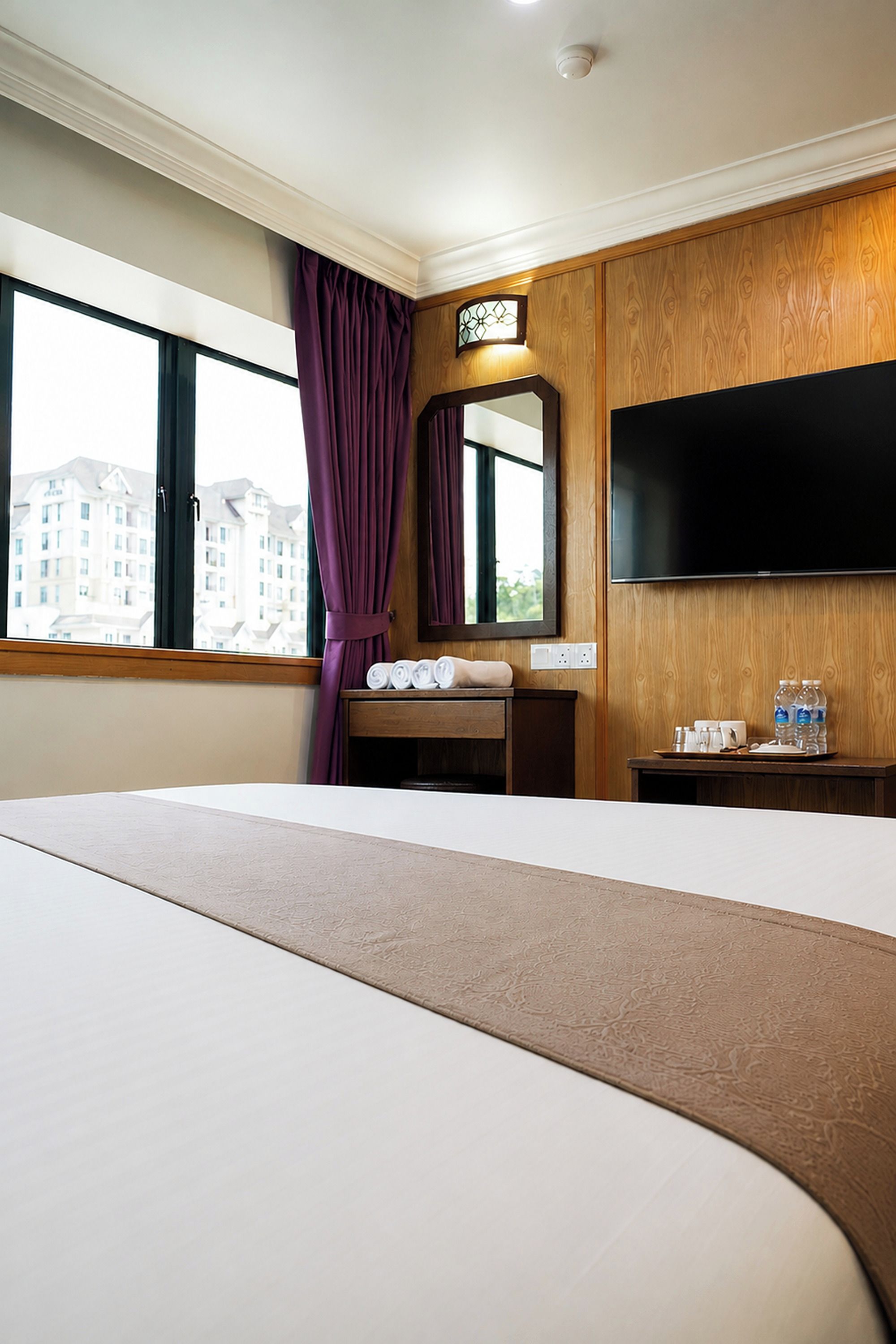 Una habitación de hotel con una cama grande, un televisor de pantalla plana montado en la pared, una ventana con cortinas moradas y vistas a edificios.