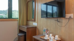 Een hotelkamer met een houten wand, een televisie boven een houten bureau, een raam met gordijnen, en een dienblad met kopjes en een fles.