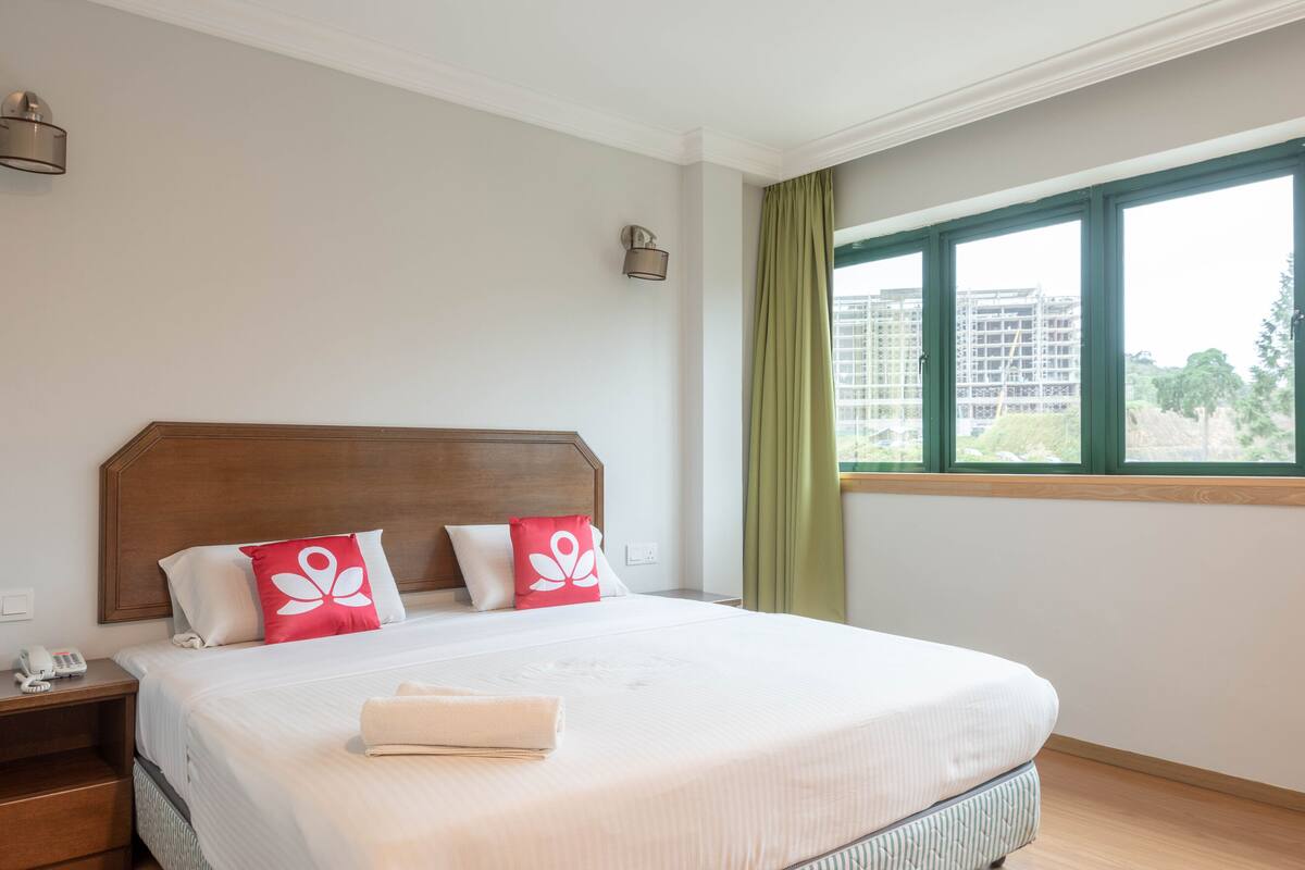 A hotel room with a large bed, a wooden headboard, a nightstand with a phone, and a window with a view of buildings and trees.