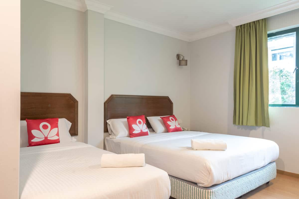 A hotel room with two beds, each with white bedding and red pillows, a wooden headboard, a small wall-mounted light, and a window with green curtains.