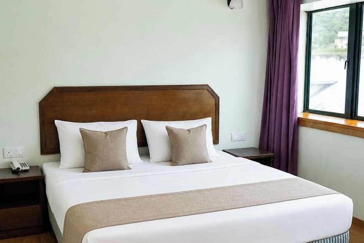 A neatly made bed with a wooden headboard, a bedside table with a phone, and a window with purple curtains.