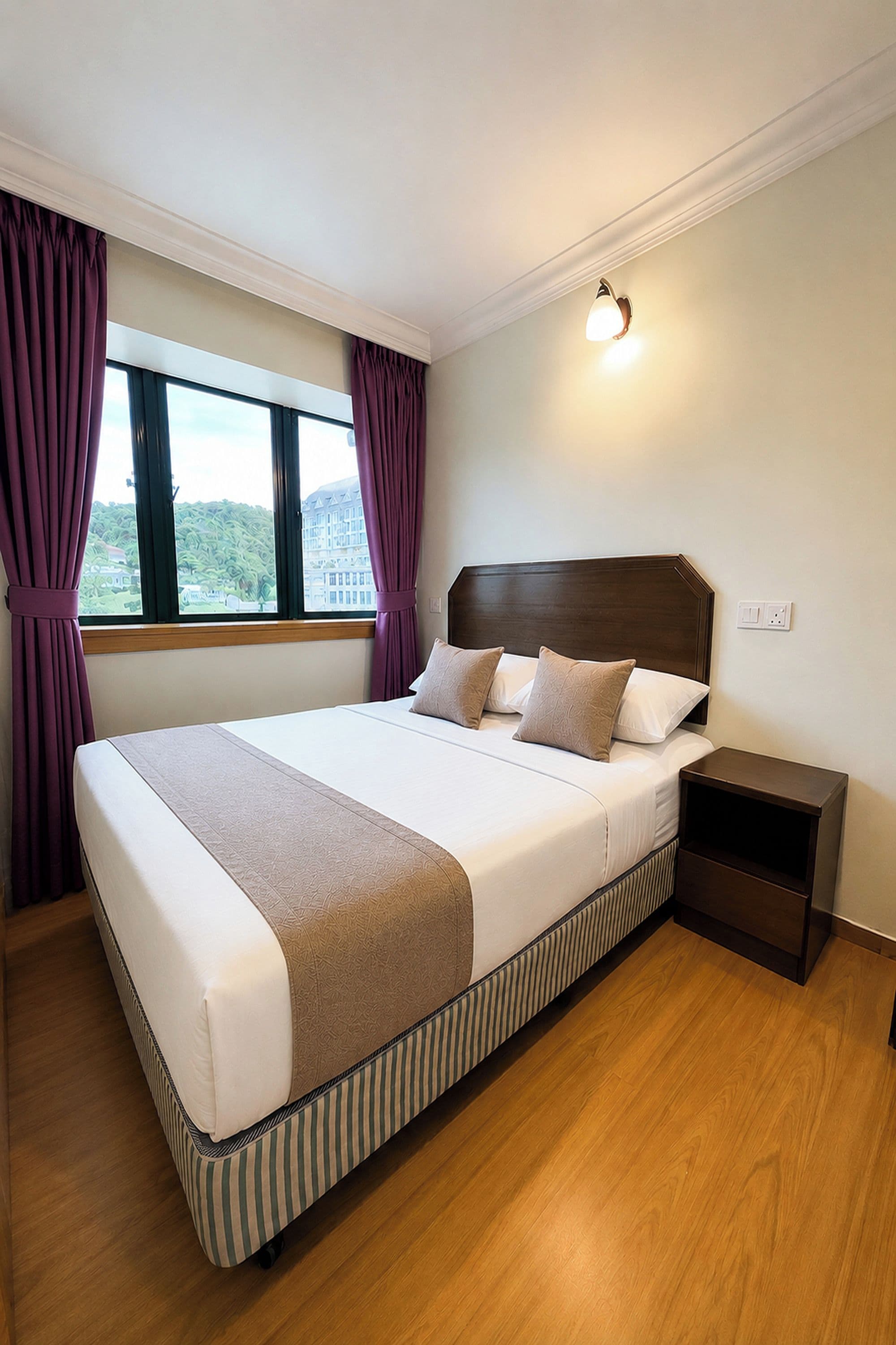 A hotel room with a large bed, a nightstand, a window with curtains, and a wall-mounted light.