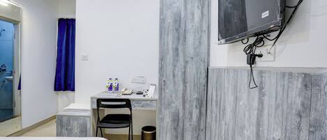 A small, modern hotel room with a bed, a desk with a chair, a TV mounted on the wall, and an air conditioning unit.