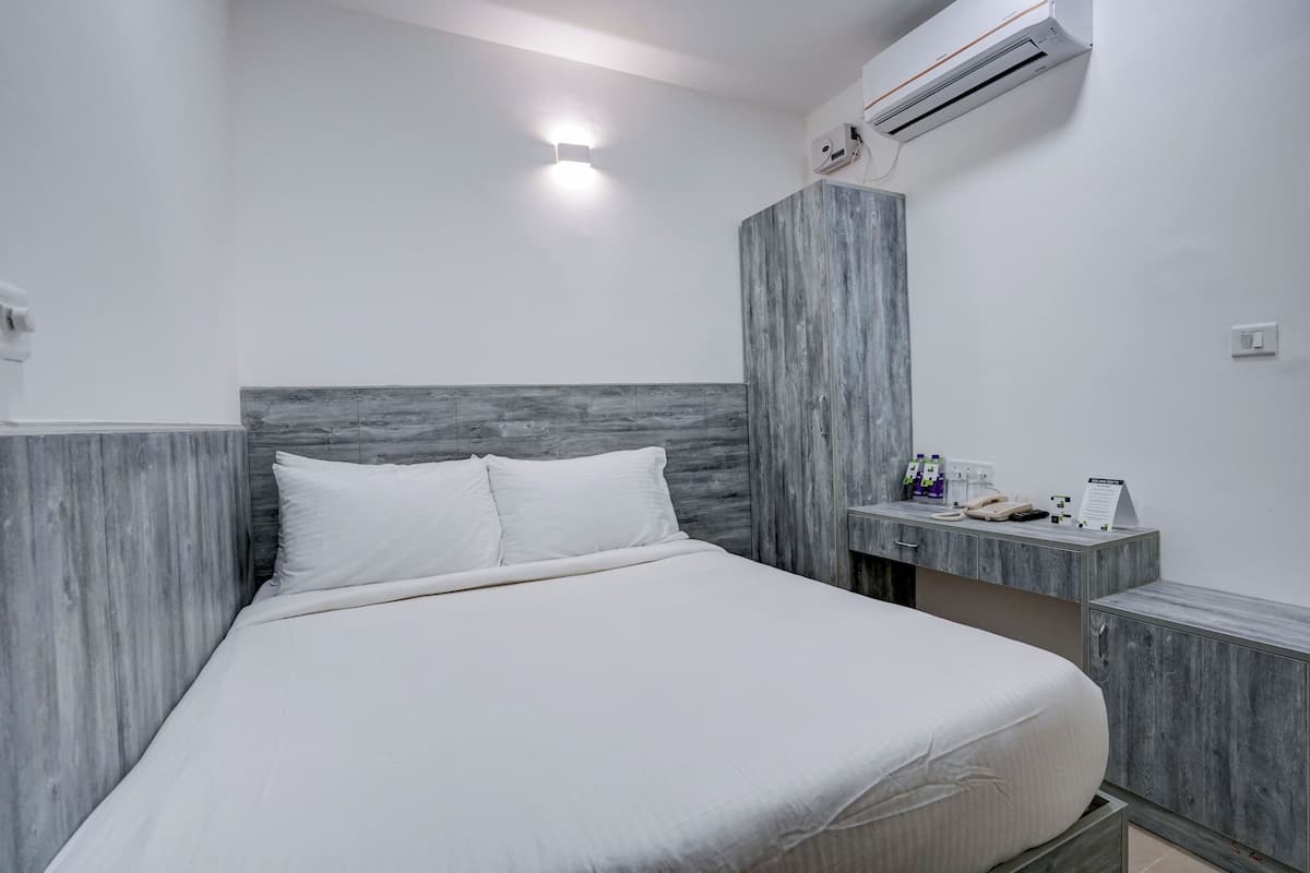 A modern hotel room with a large bed, a wooden headboard, a bedside table with toiletries, and an air conditioning unit.