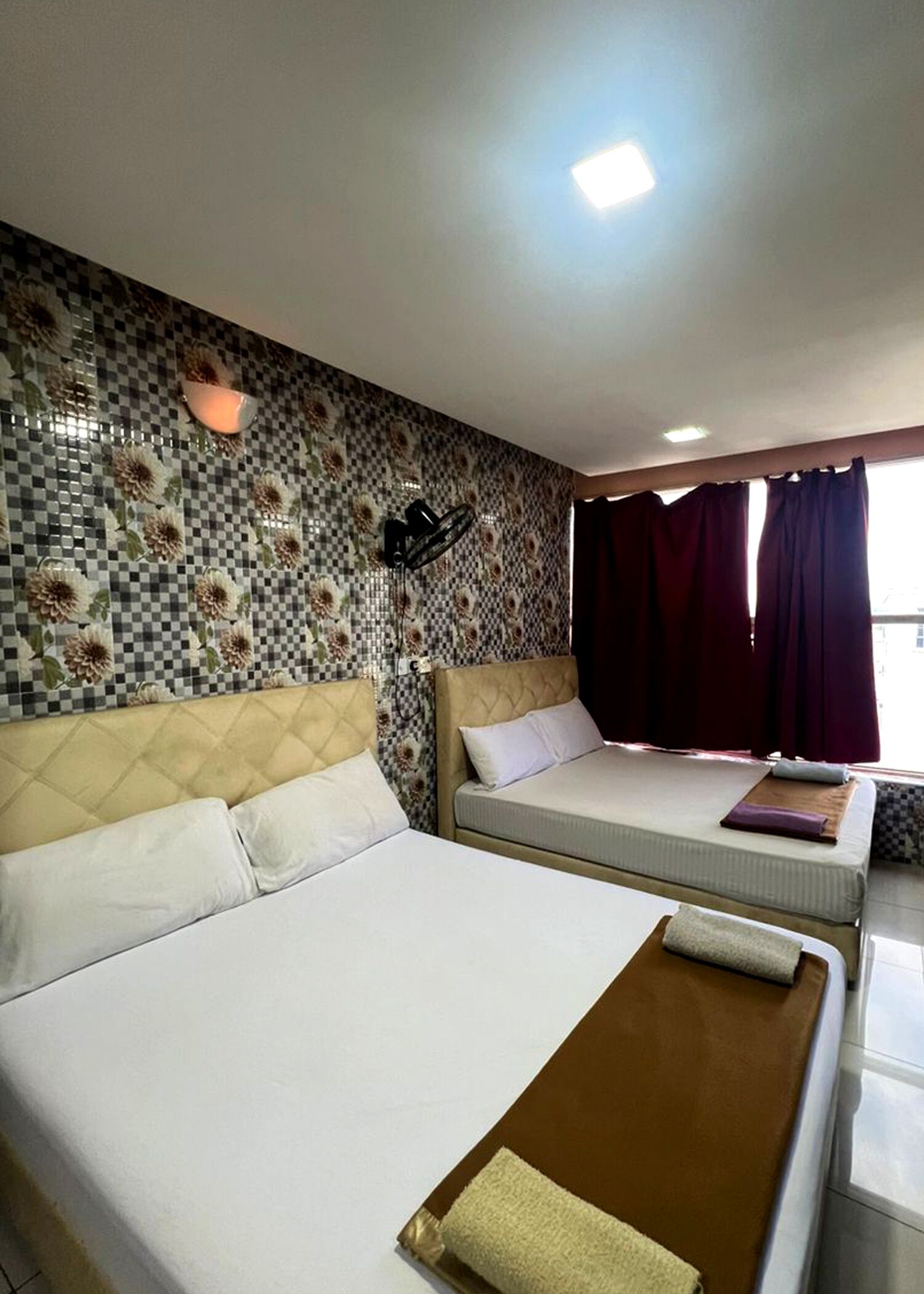 Habitación de hotel con dos camas, un papel tapiz floral a cuadros, un ventilador de techo y una ventana con cortinas.