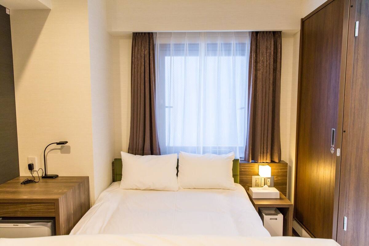 A hotel room with a bed, a nightstand with a lamp, a wooden wardrobe, and a window with curtains.