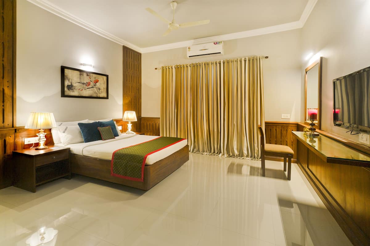 Quarto de hotel com cama, mesas de cabeceira, escrivaninha, cadeira, espelho, ventoinha no teto e janela com cortinas.