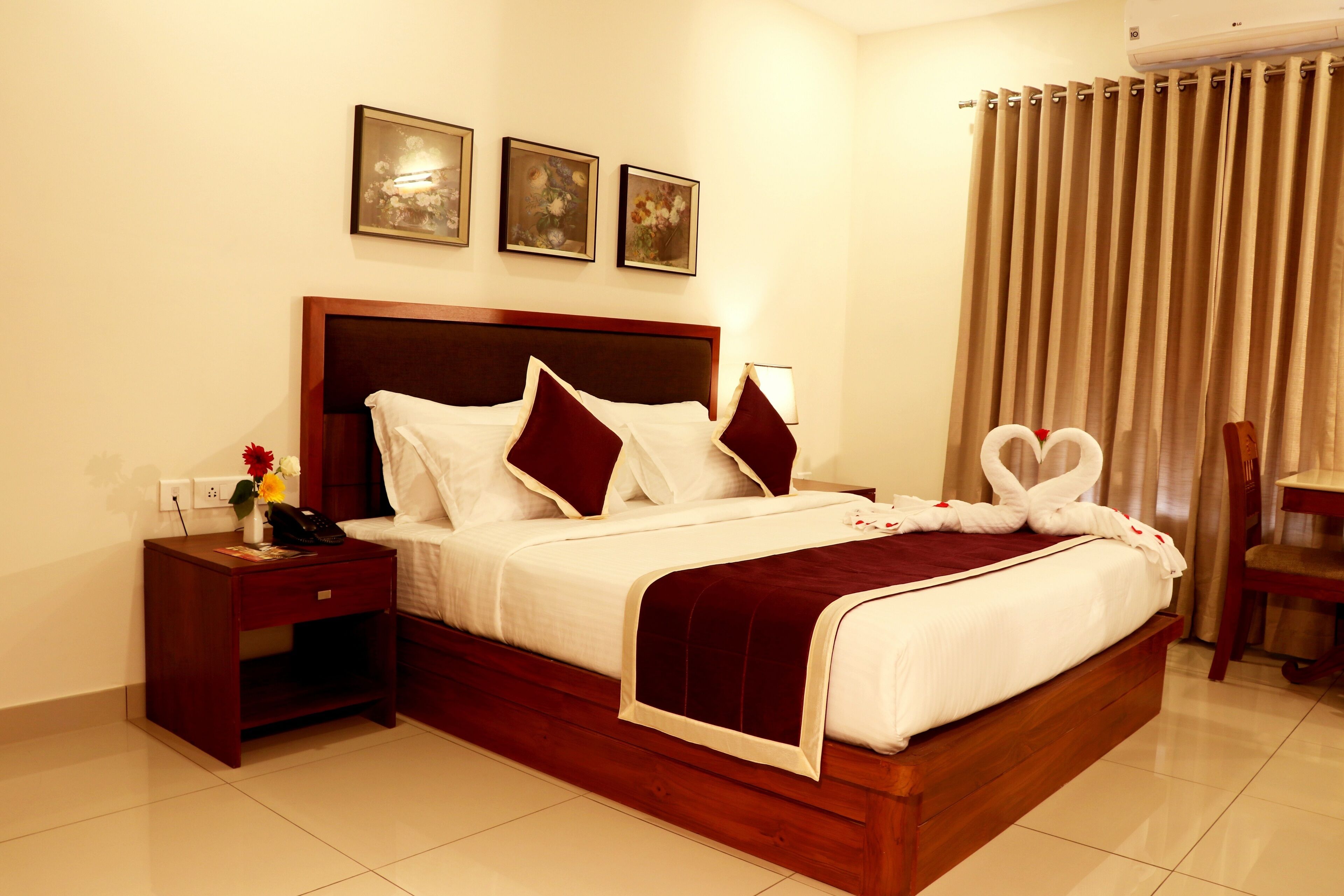 A neatly made bed with white linens and red pillows, a wooden nightstand with a phone and a vase of flowers, and a chair with a table beside the bed.