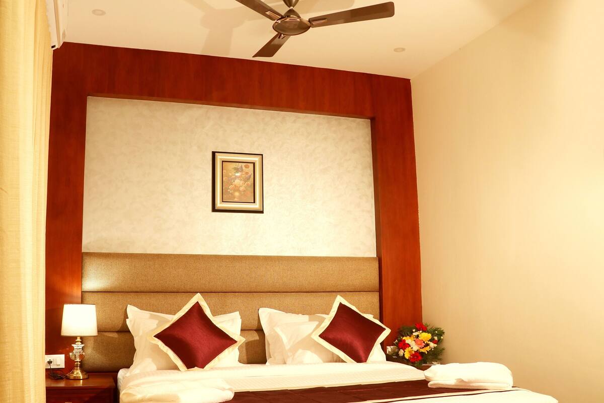 Quarto de hotel com uma cama grande, um ventilador de teto, uma mesa de cabeceira com abajur e um quadro na parede.
