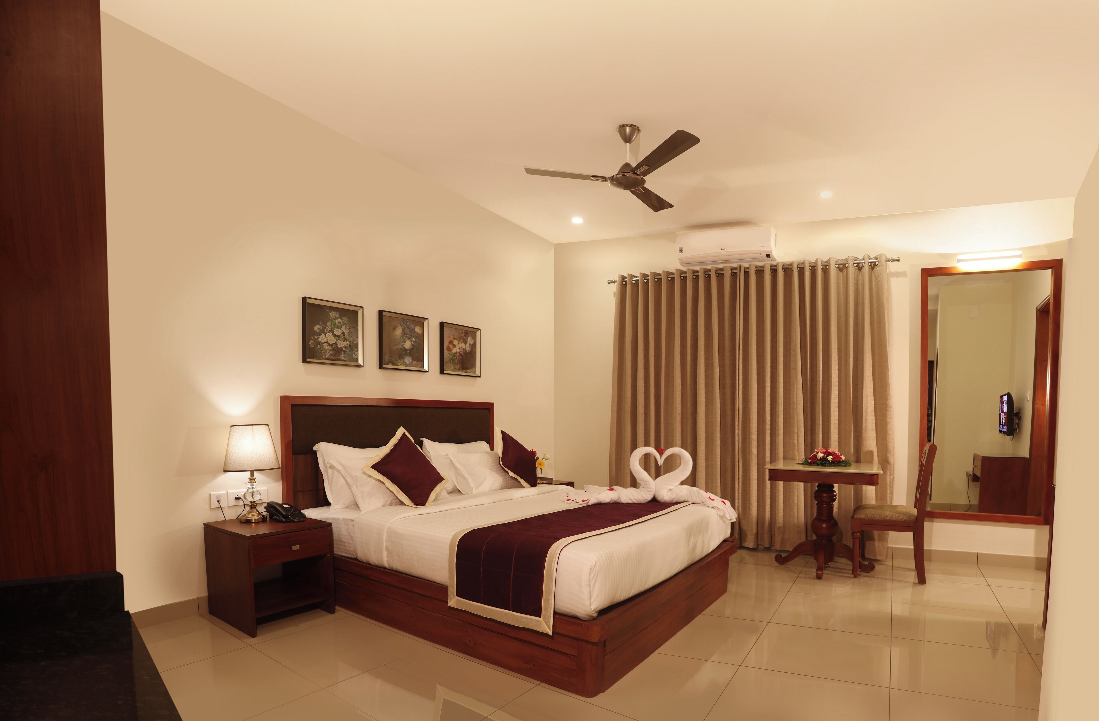 A hotel room with a large bed, a bedside table with a lamp, a ceiling fan, and a small table with a flower arrangement.