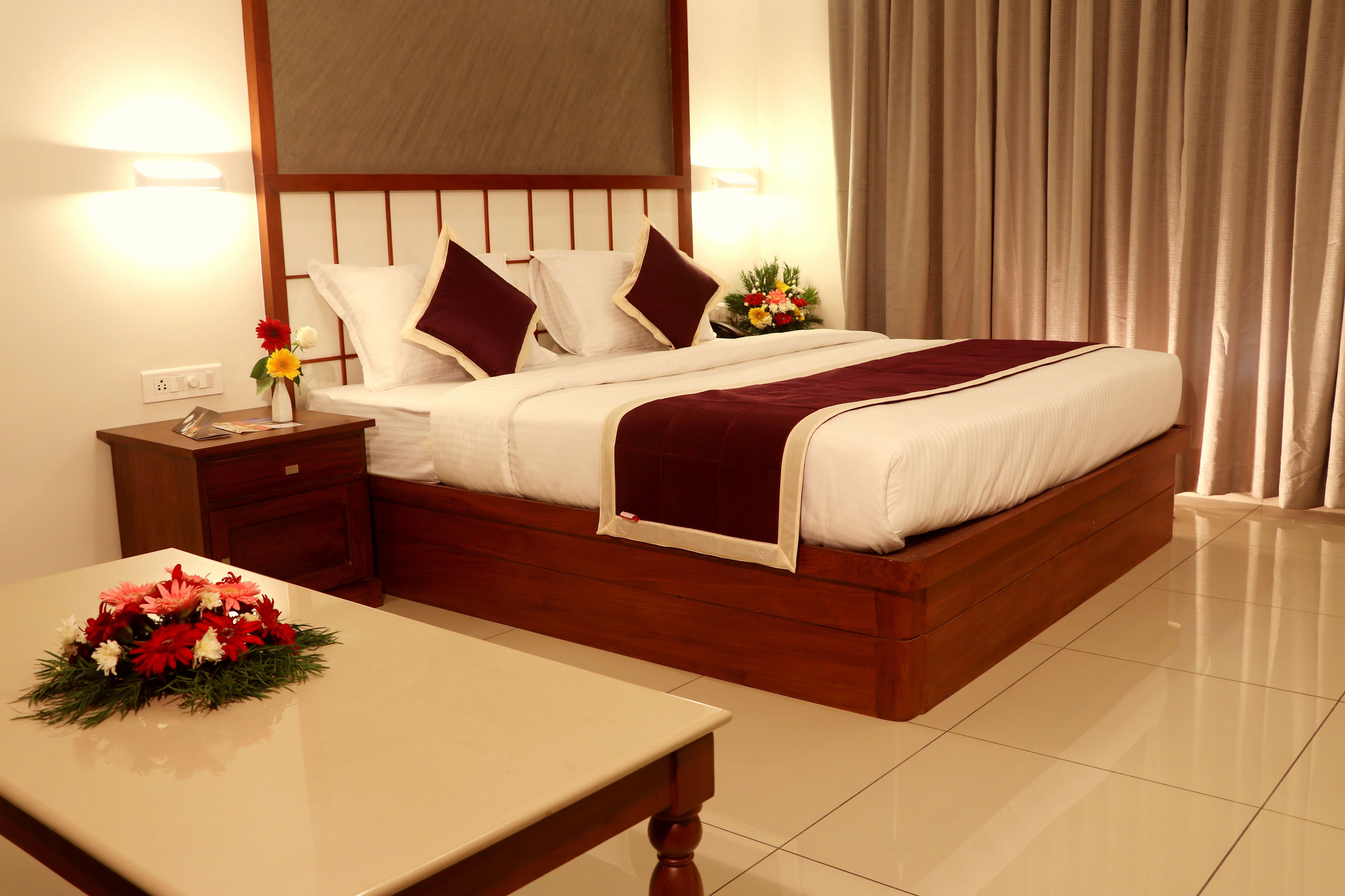 A hotel room with a large bed, wooden headboard, bedside tables, and a small table with a floral arrangement.