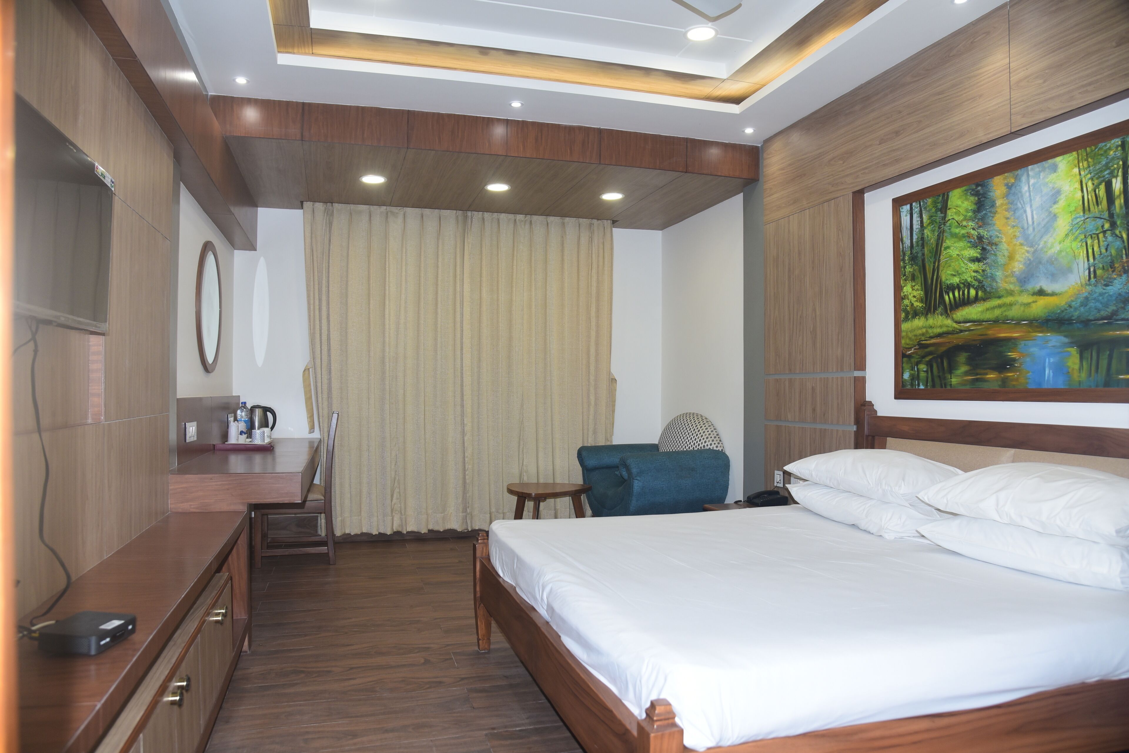 Habitación de hotel con una cama grande, un escritorio con silla, un televisor y un cuadro de un paisaje boscoso.