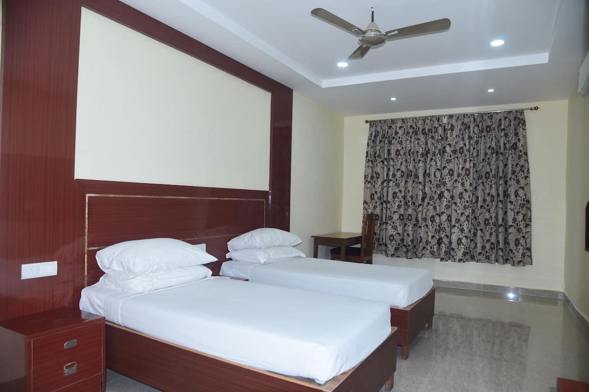 Quarto de hotel com duas camas de madeira, um ventilador de teto, uma mesa pequena e uma cortina estampada.