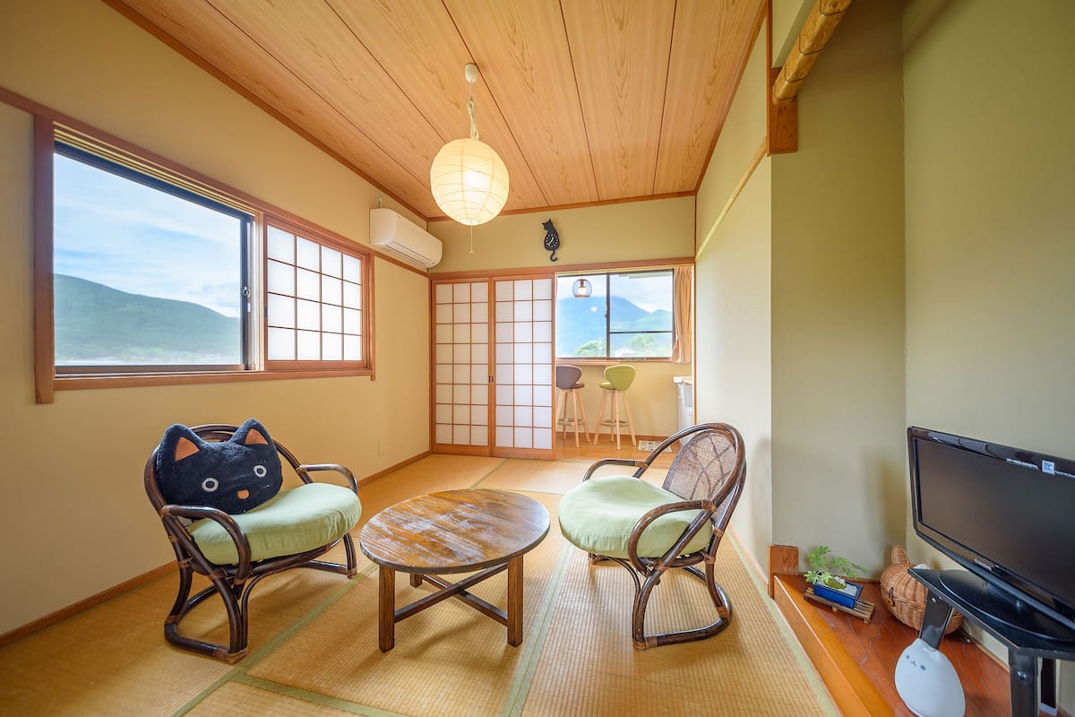 Ein traditioneller japanischer Raum mit Tatami-Böden, einem niedrigen Tisch, zwei katzenförmigen Stühlen, einem Fernseher und Blick ins Freie.