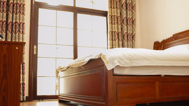 Een houten bed met een witte matras en hoofdbord, geplaatst naast een houten kastje en een groot raam met uitzicht.