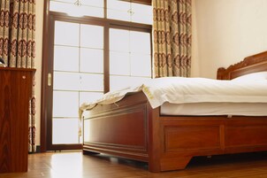 A wooden bed with a white mattress and a headboard, positioned next to a wooden cabinet and a large window with a view of the outside.