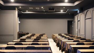 Un salón de conferencias con filas de mesas y sillas, un atril y una gran pared gris.