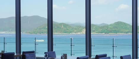 A restaurant with large windows offering a view of the sea and hills. Tables are set with chairs, and the Stanford Hotel & Resort Tongyeong logo is visible.