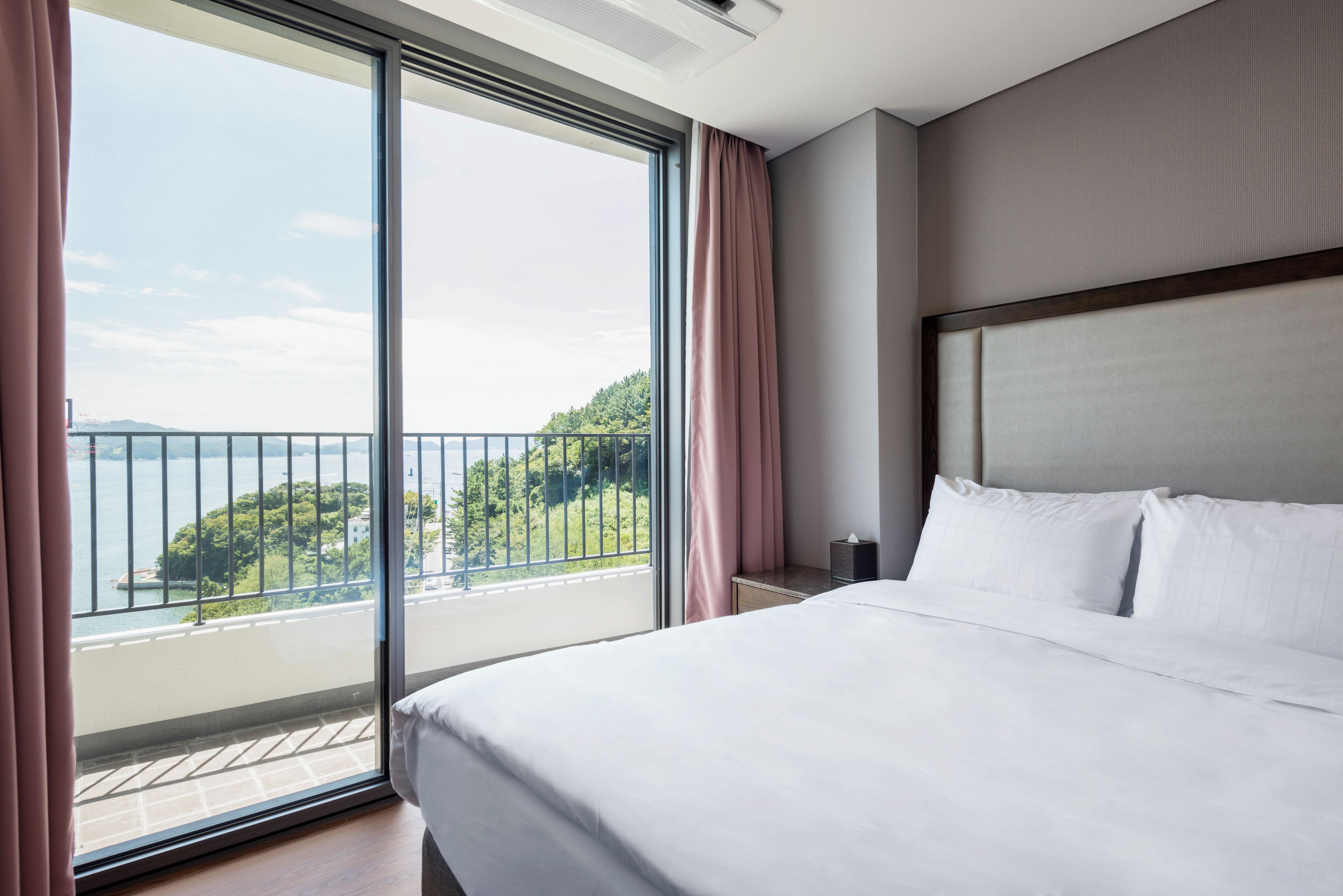 A hotel room with a large bed, a nightstand, a window with curtains, and a view of a body of water and greenery.