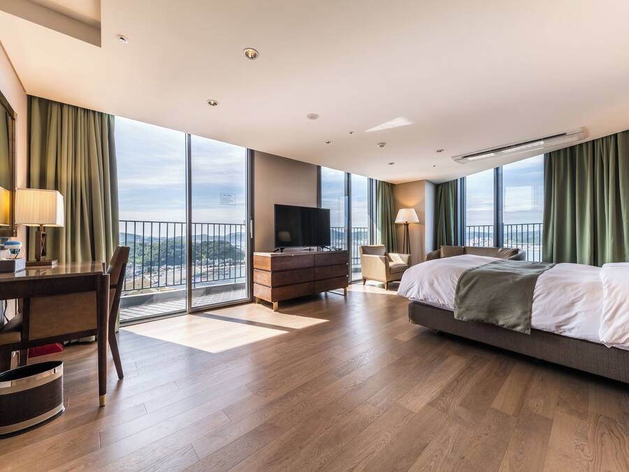 Habitación de hotel con una cama grande, un escritorio con silla, televisión y vista a la ciudad a través de amplias ventanas.