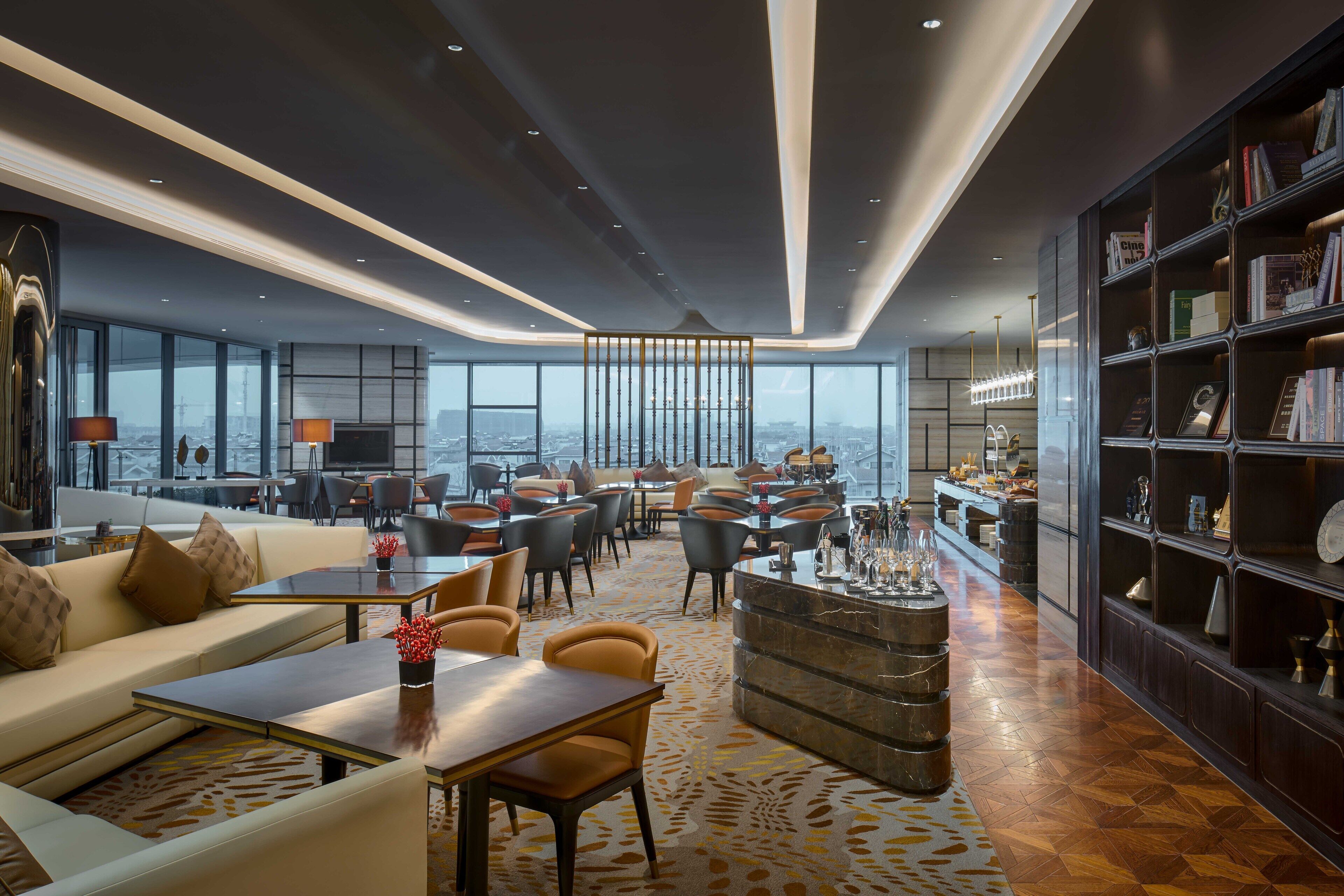 Una hall d'albergo moderna con soffitto alto, ampie finestre che offrono vista sulla città, un'area bar centrale e un salotto con divani e tavolini.