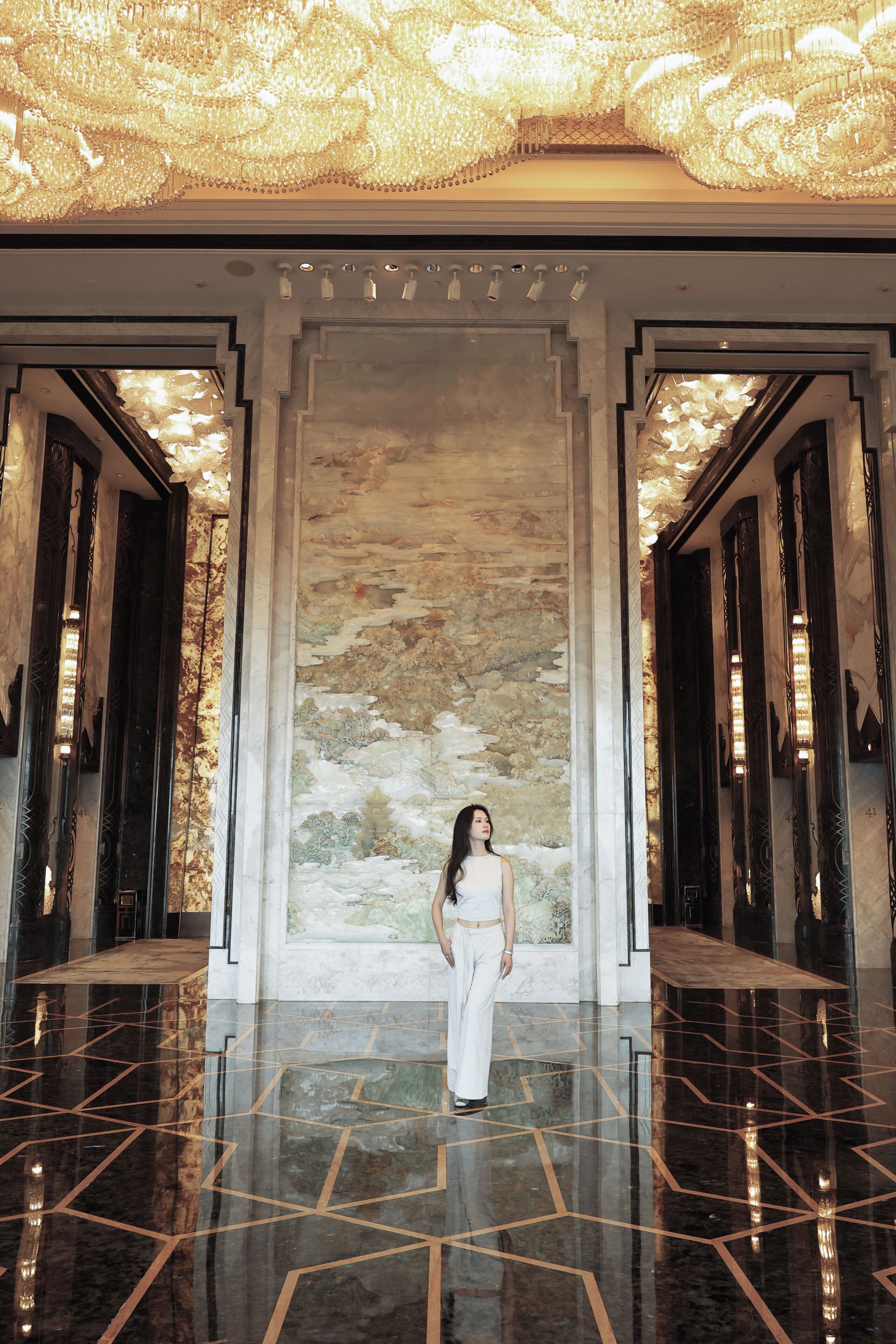 A woman standing in a hallway with a large mural, polished floors, and ornate ceiling lights.