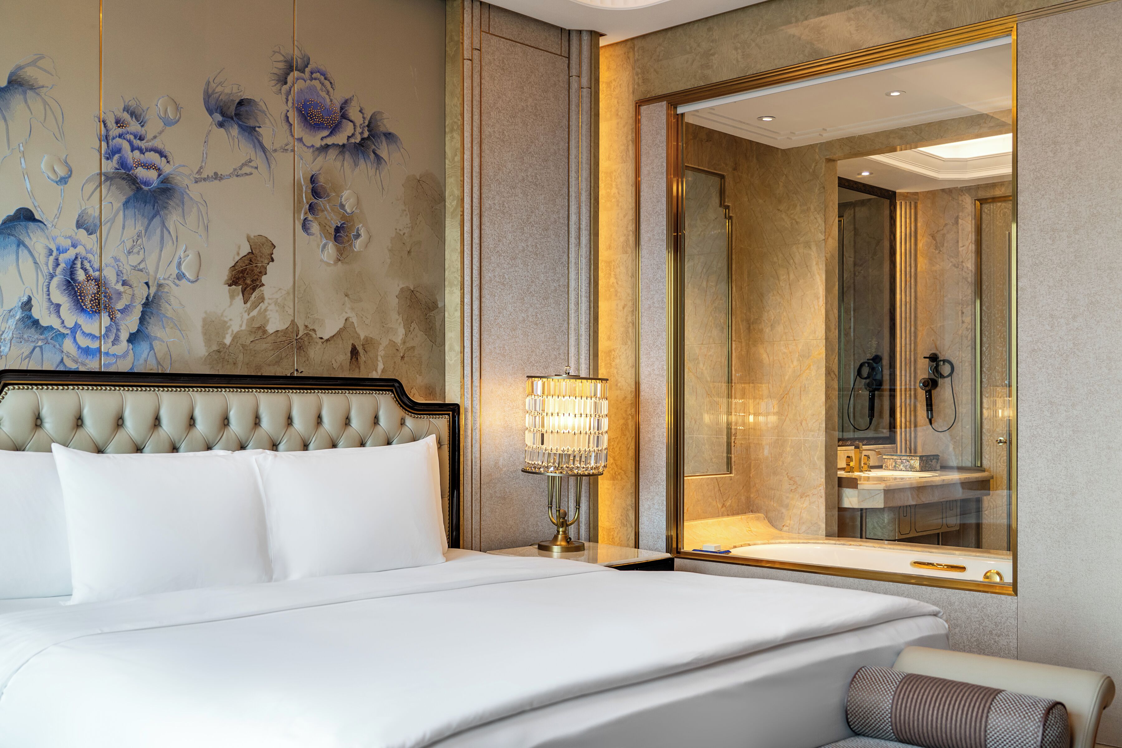 Een slaapkamer met een groot bed, een nachtkastje met een lamp, een spiegel en een wand met bloemwerk.