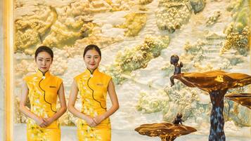 Dos personas vestidas de amarillo de pie frente a un mural con un paisaje montañoso y una mesa con una estatua.