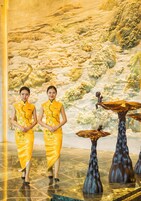 Dos personas vestidas de amarillo de pie frente a un mural con un paisaje montañoso y una mesa con una estatua.