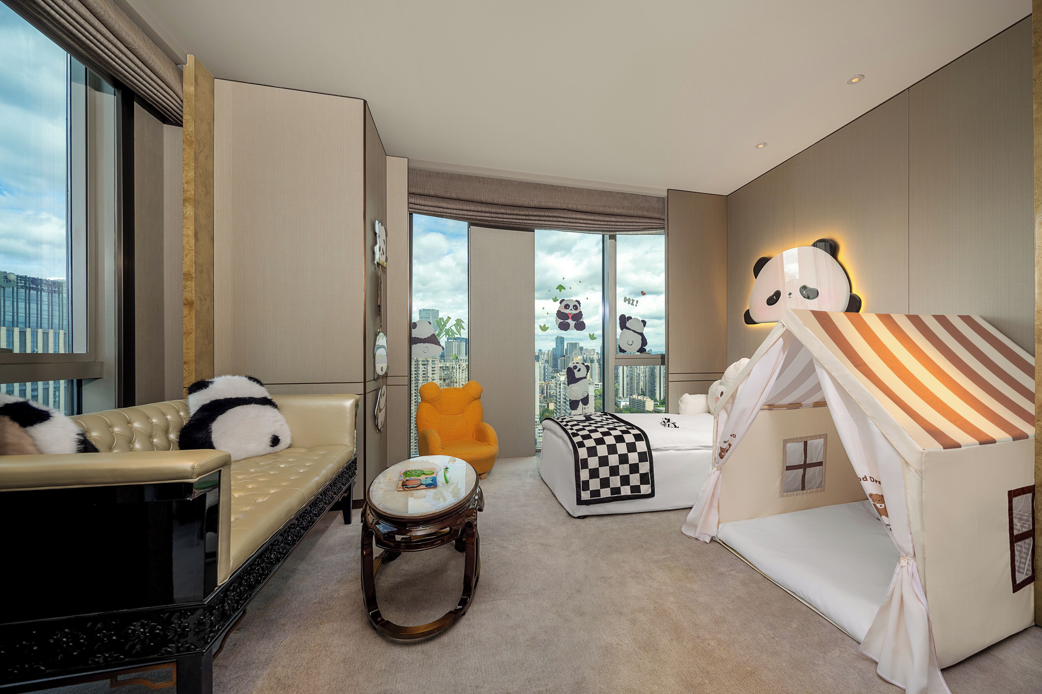 A hotel room with a sofa, a small round table, a bed with a striped canopy, a panda-themed play tent, and a large window with a city view.