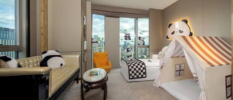 Quarto de hotel com sofá, mesinha redonda, cama com dossel listrado, tenda infantil com tema de panda e uma janela grande com vista para a cidade.