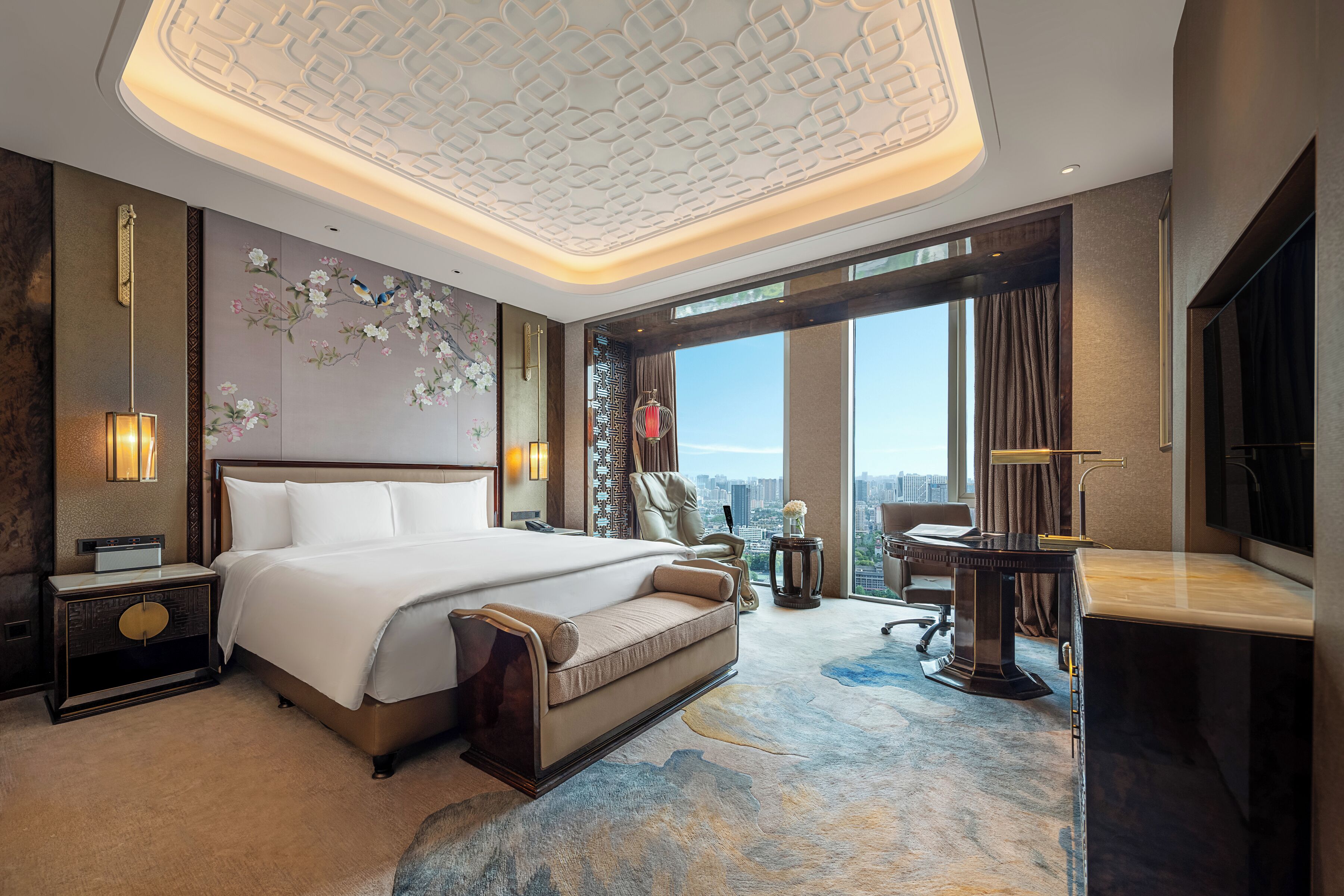 A hotel room with a large bed, a desk, a chair, a TV, and a view of the city.