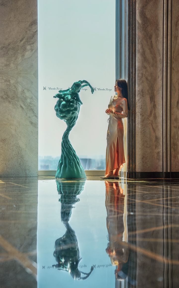 Una persona de pie junto a una escultura en un vestíbulo moderno con piso reflectante y ventanas amplias.