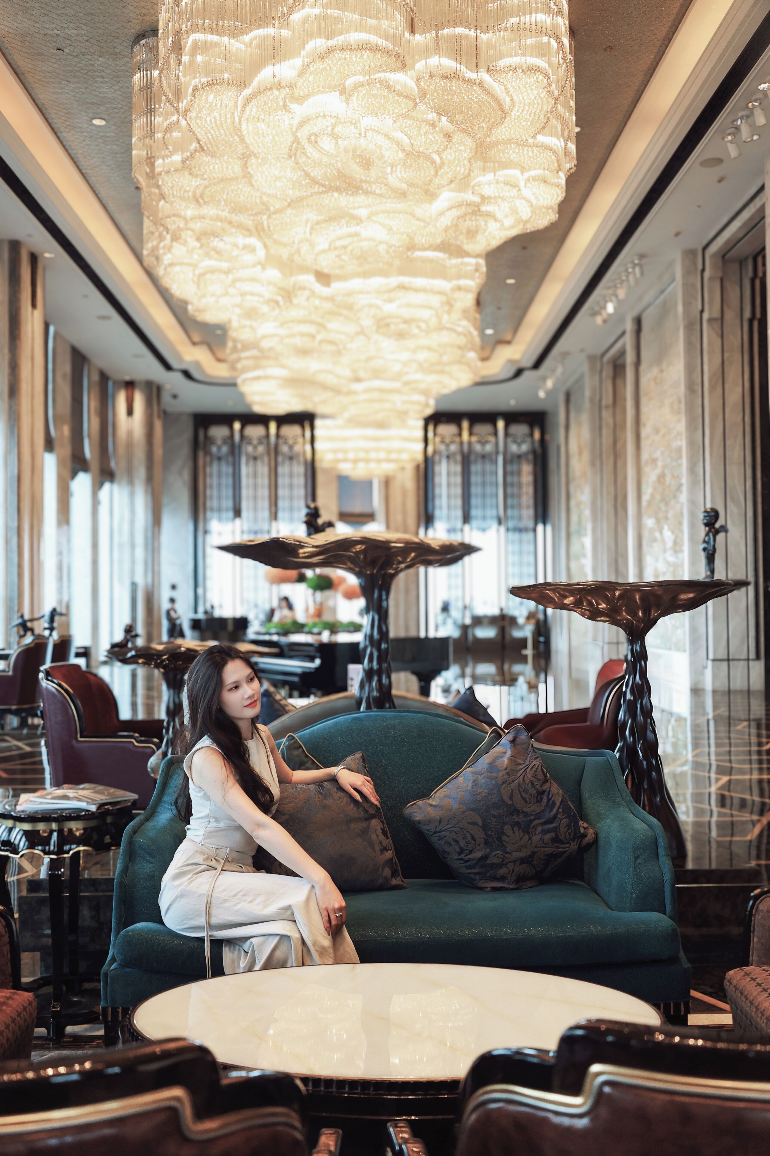 A woman seated on a teal sofa in a modern hotel lobby with a large chandelier, a piano, and a dining area in the background.