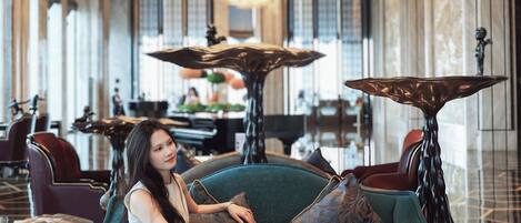 Una mujer sentada en un sofá color turquesa en un moderno vestíbulo de hotel, con una lámpara de araña grande, un piano y un área de comedor al fondo.