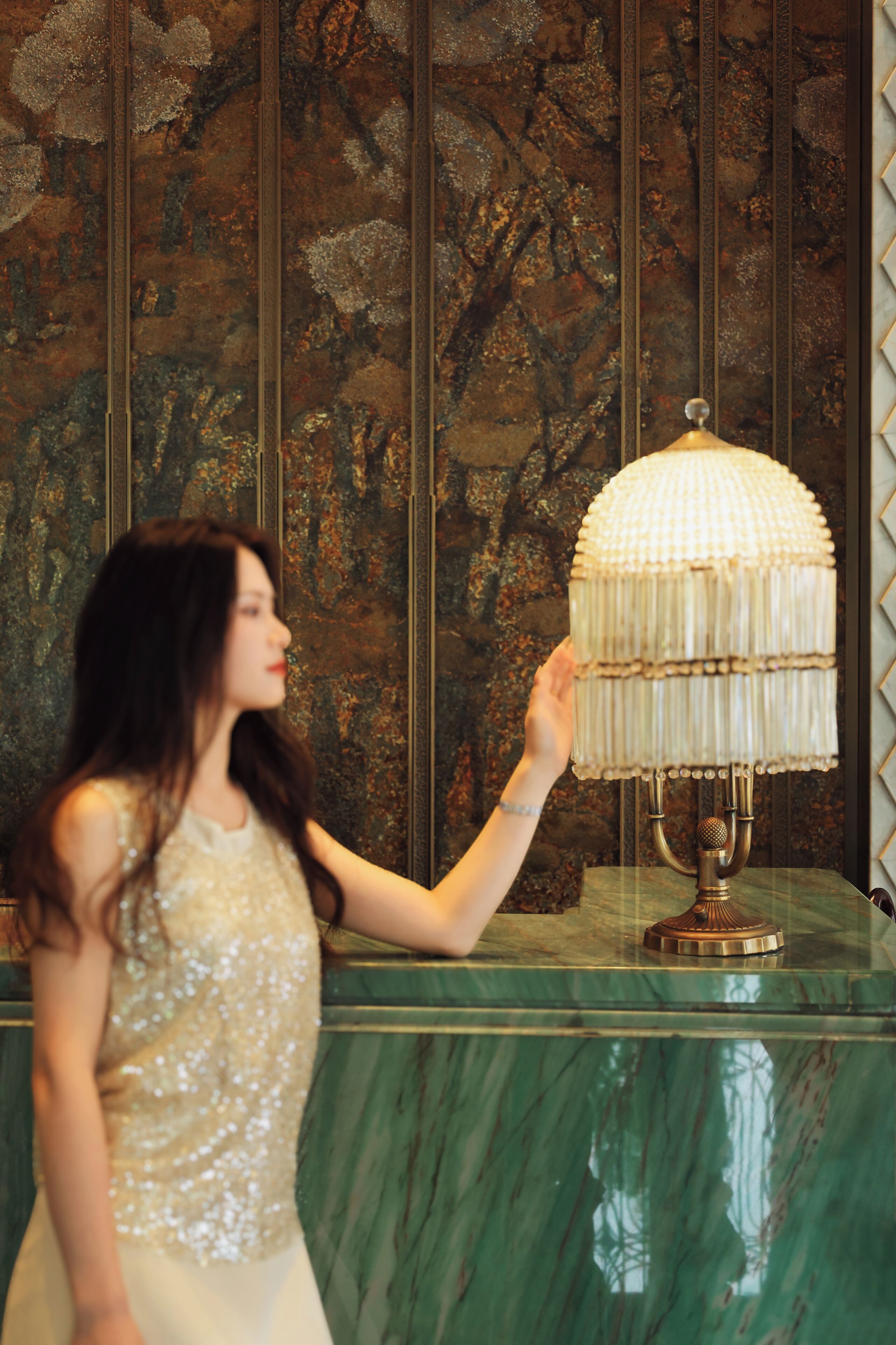 A woman in a sequined top standing next to a green marble table with a decorative lamp.
