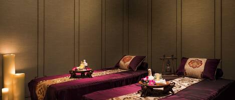 A room with two beds, each with a purple bedspread and a tray of toiletries. There are candles on the floor beside the beds, and a wall-mounted lamp is visible.