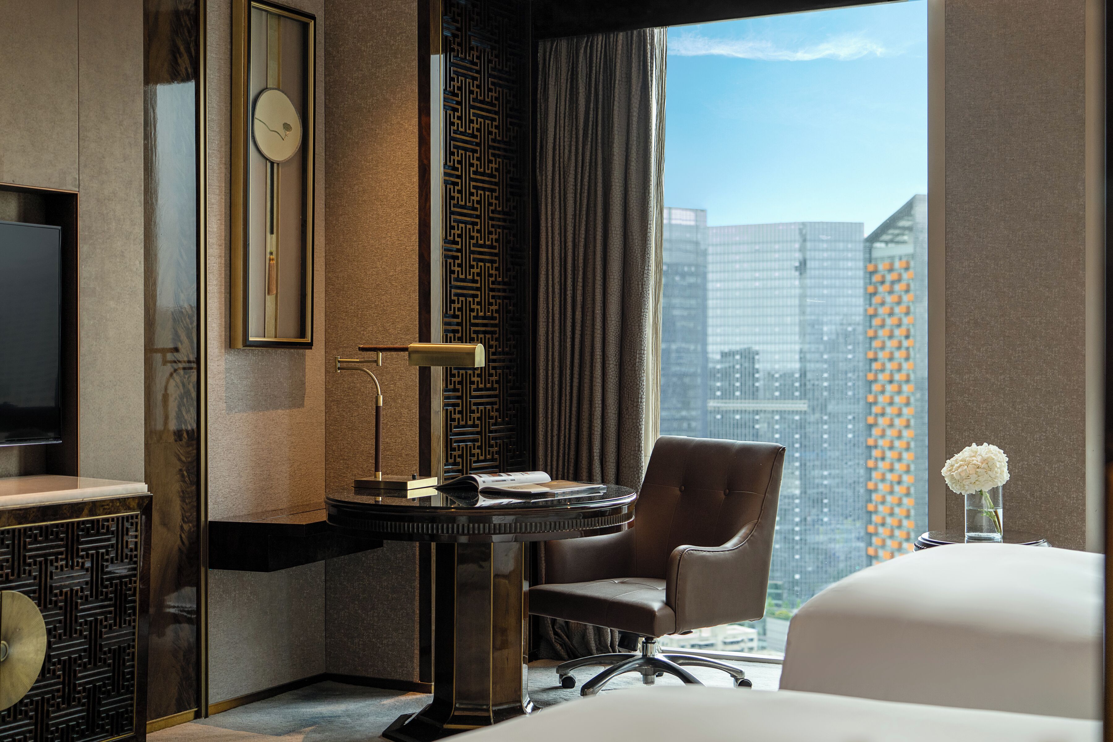 A hotel room with a desk, chair, TV, and a view of the cityscape.