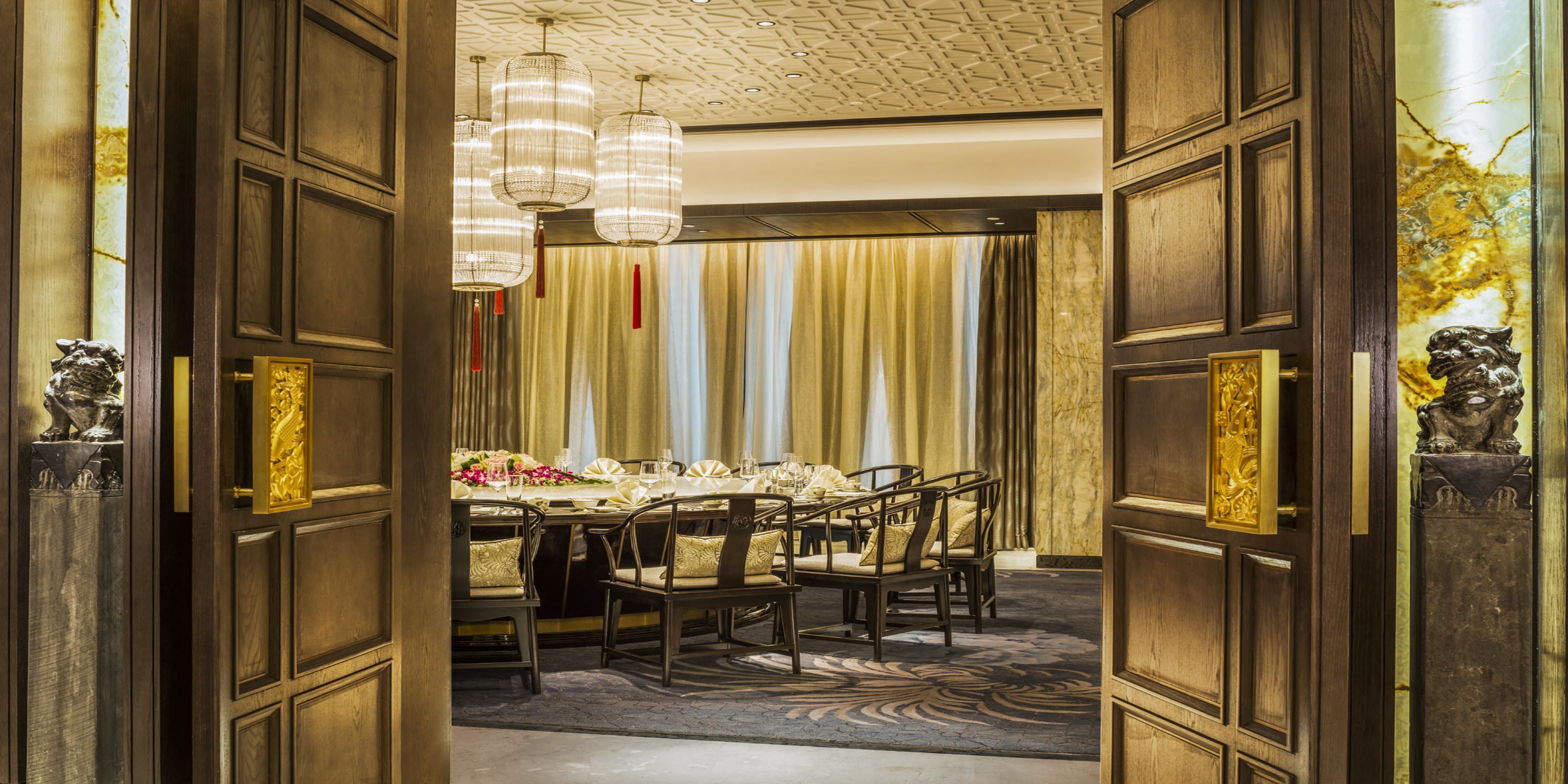 A formal dining room with a long table set for multiple guests, flanked by ornate wooden doors with gold accents.
