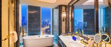 A luxurious bathroom with a large bathtub, dual sinks, a vanity with a mirror, and a city view through the windows.