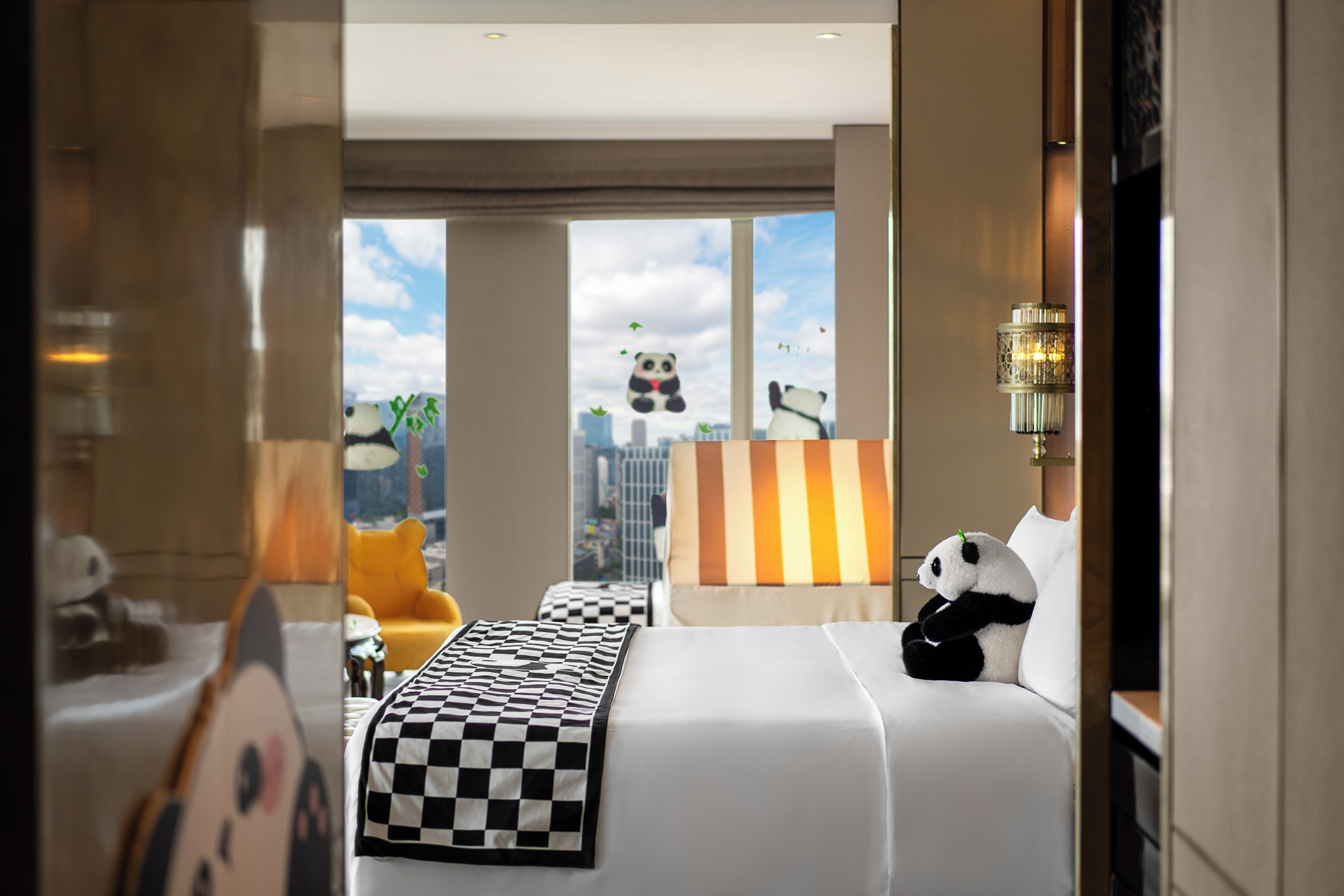 A hotel room with a bed, a window with a city view, a lamp, and a panda plush toy.