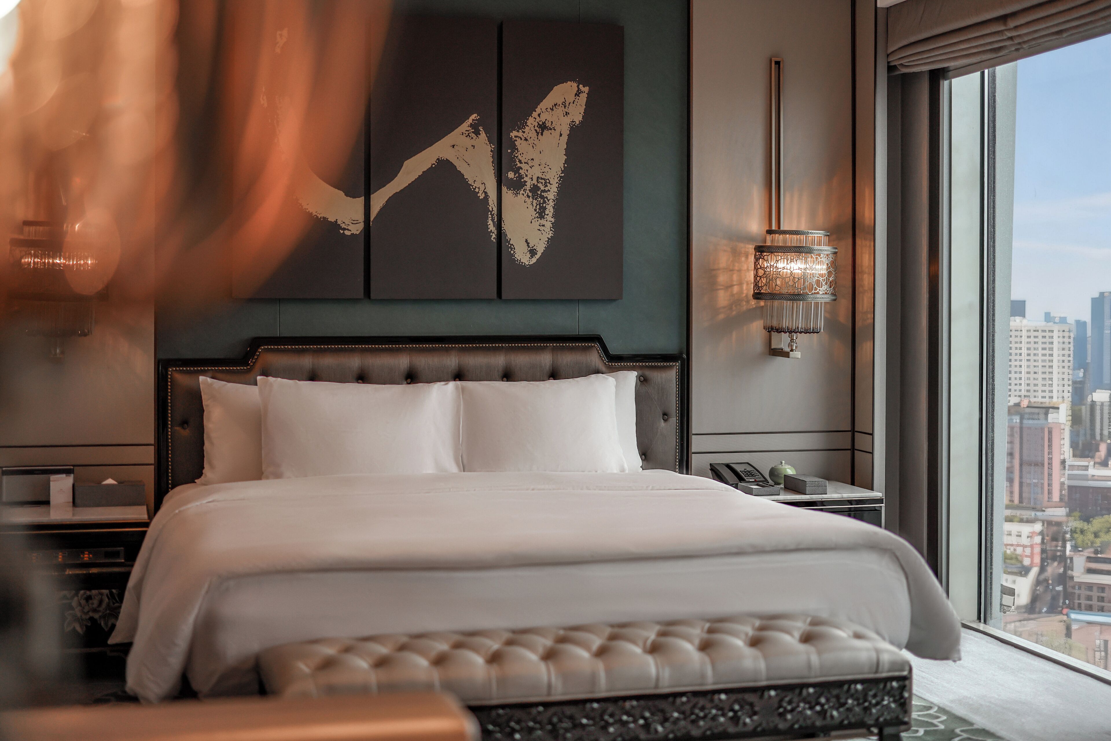 A modern hotel room with a large bed, a city view, and a decorative wall piece.