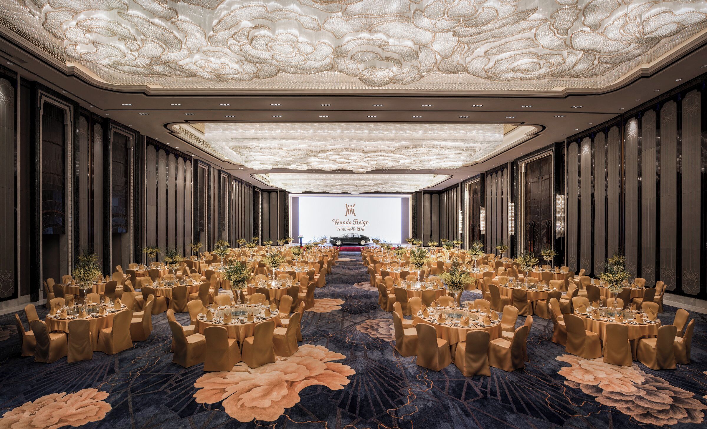 A banquet hall with round tables, chairs, and a stage with a car display.