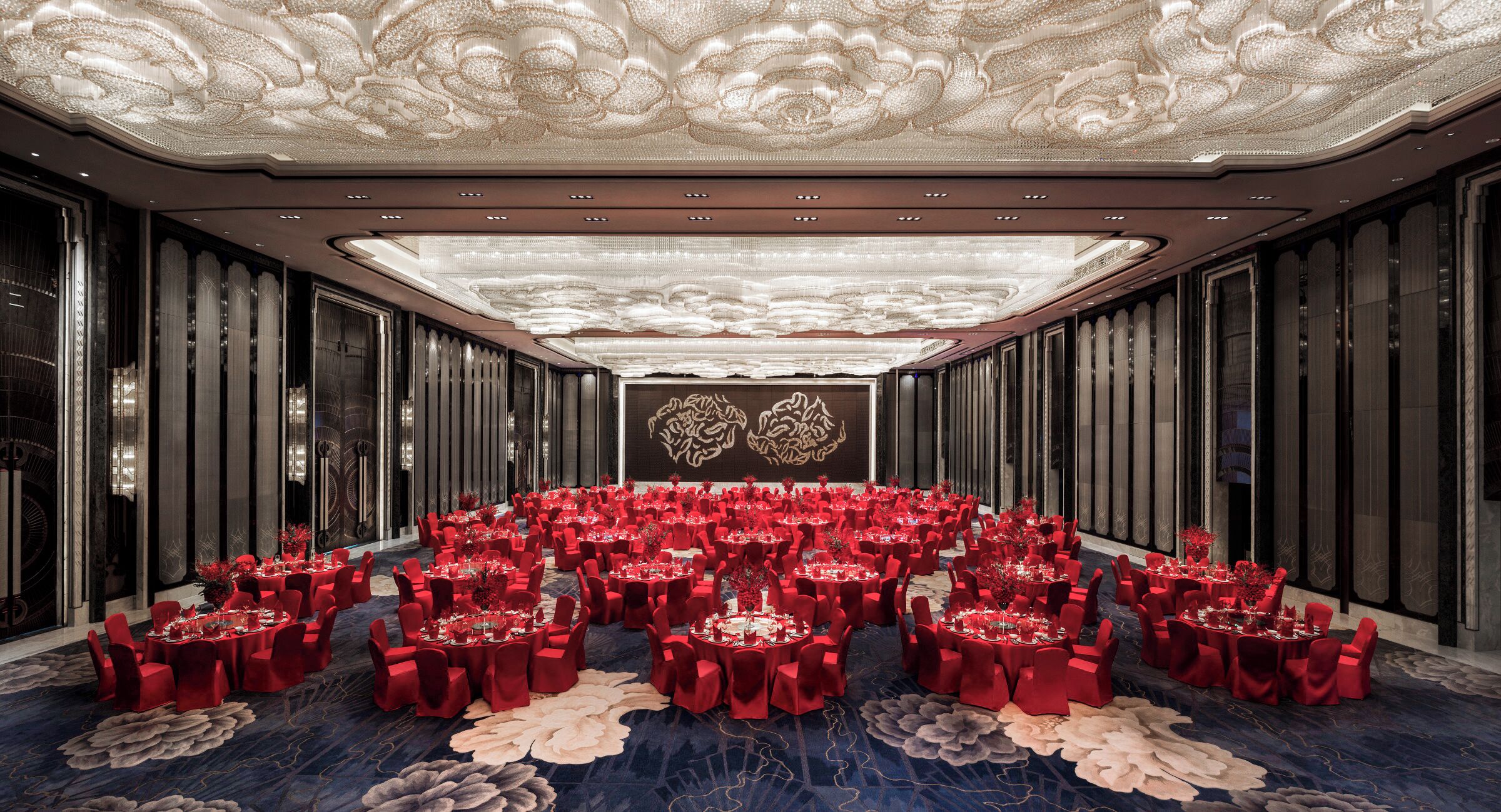 A large banquet hall with round tables, red chairs, and a decorative ceiling.