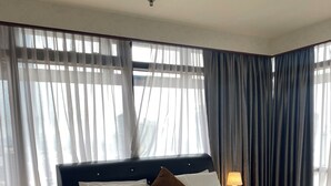 A neatly made bed with white linens and a brown pillow, a bedside lamp, and a view through sheer curtains. - Eastern Suites at Times Square KL (Kuala Lumpur)