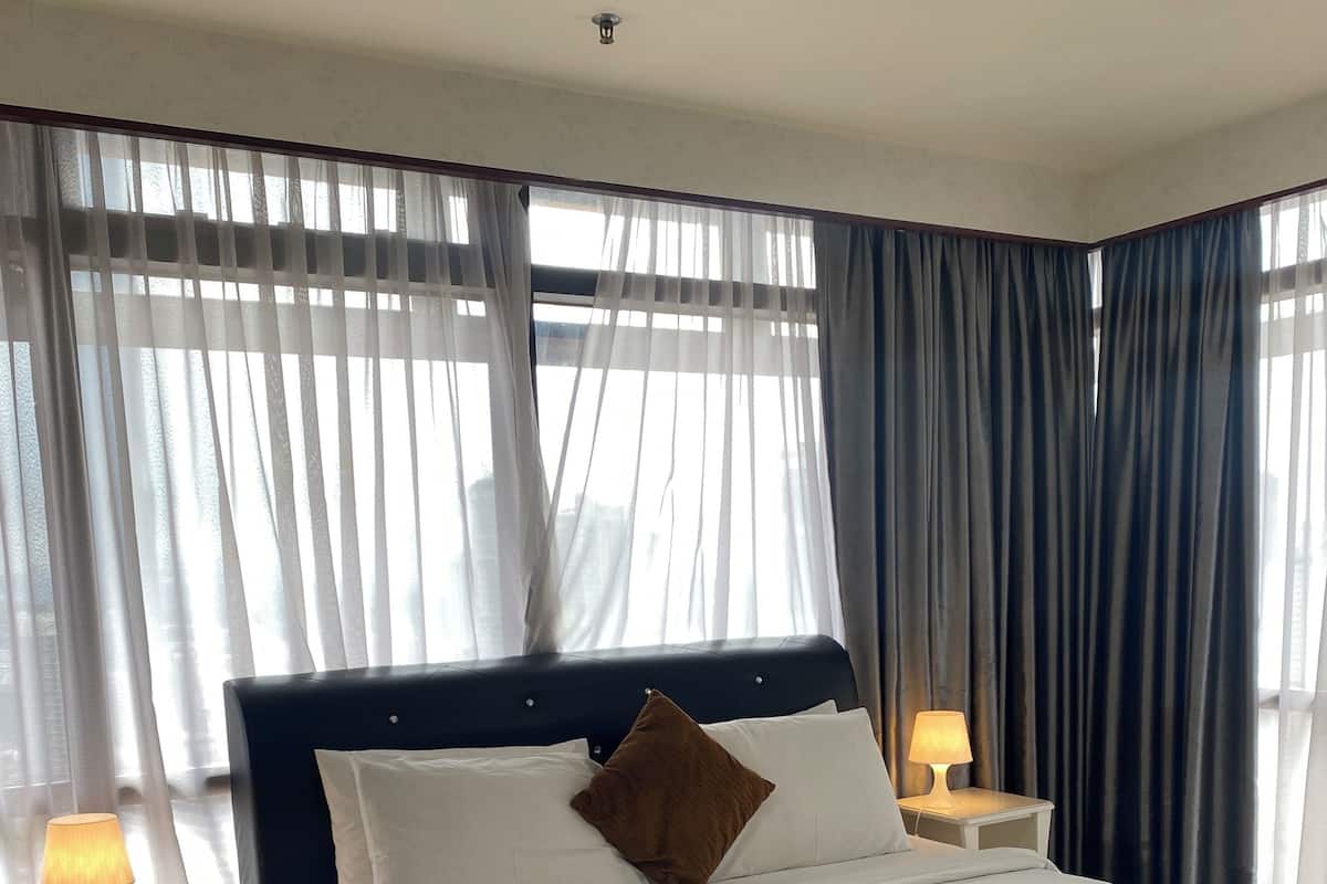 A neatly made bed with white linens and a brown pillow, a bedside lamp, and a view through sheer curtains.